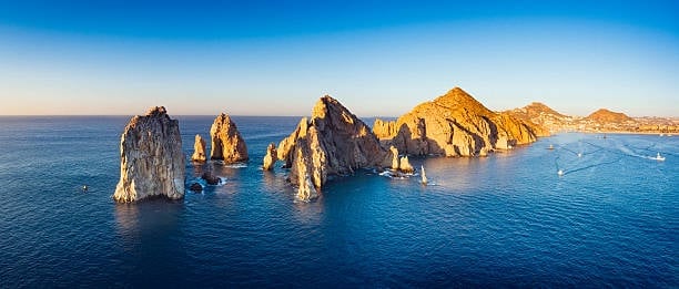 Dramatic rocky peaks and sea stacks rising from bright blue ocean water under clear sky, with sailboats visible in the distance