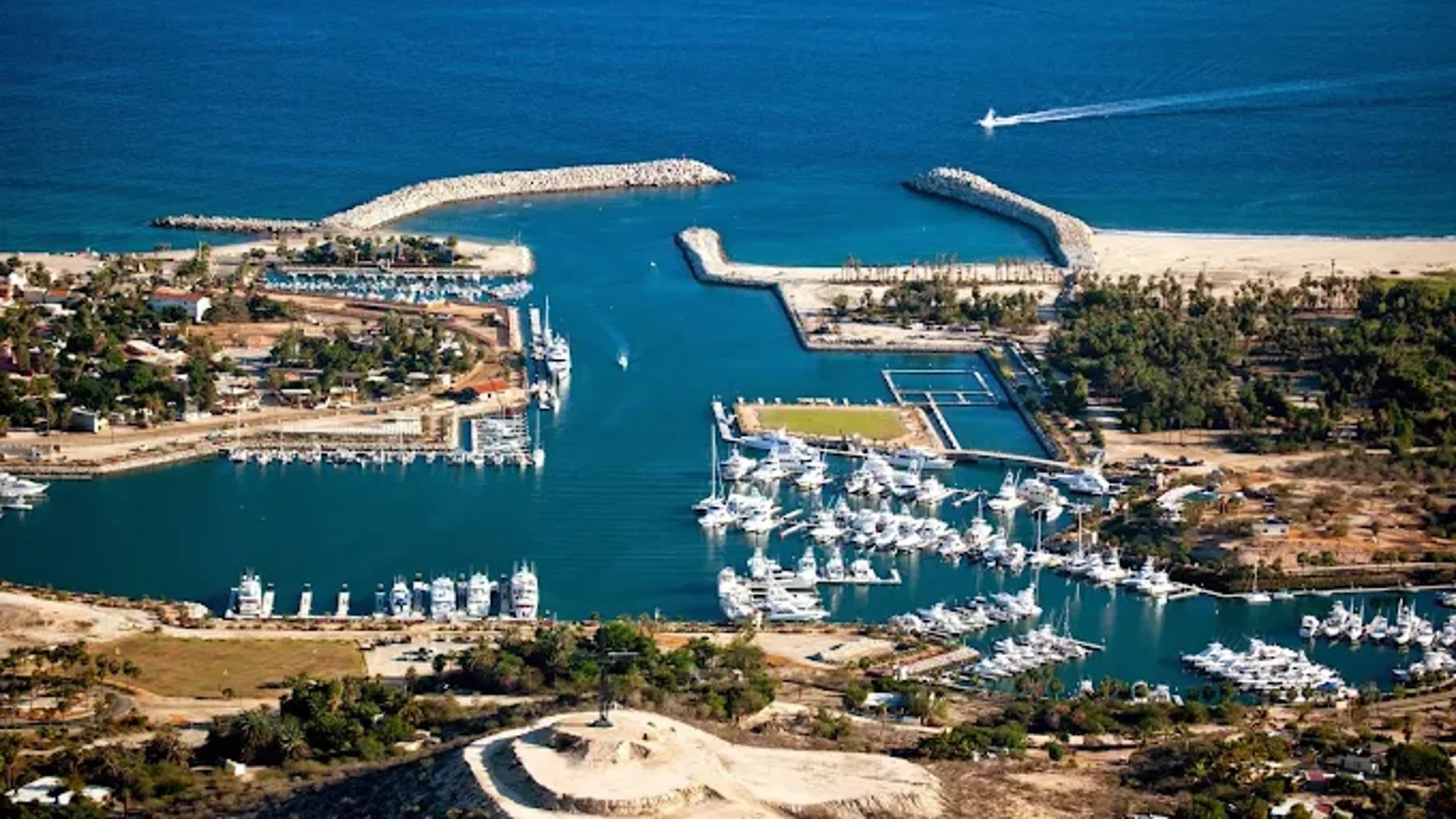 Aerial view of a coastal marina with white boats docked in turquoise water, surrounded by sandy beaches and green vegetation