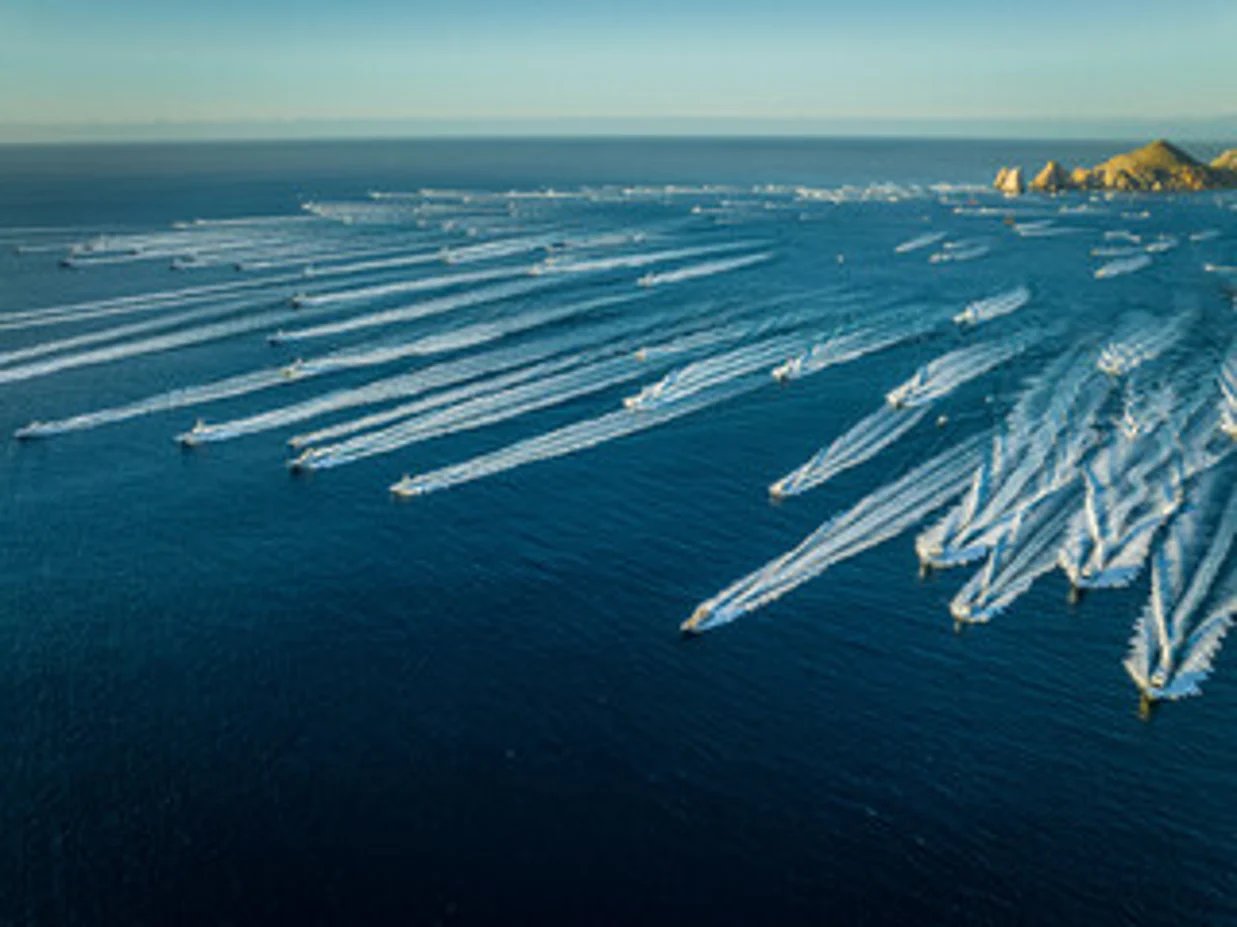 Aerial view of multiple speedboats creating white wake trails in blue ocean water near rocky coastal cliffs