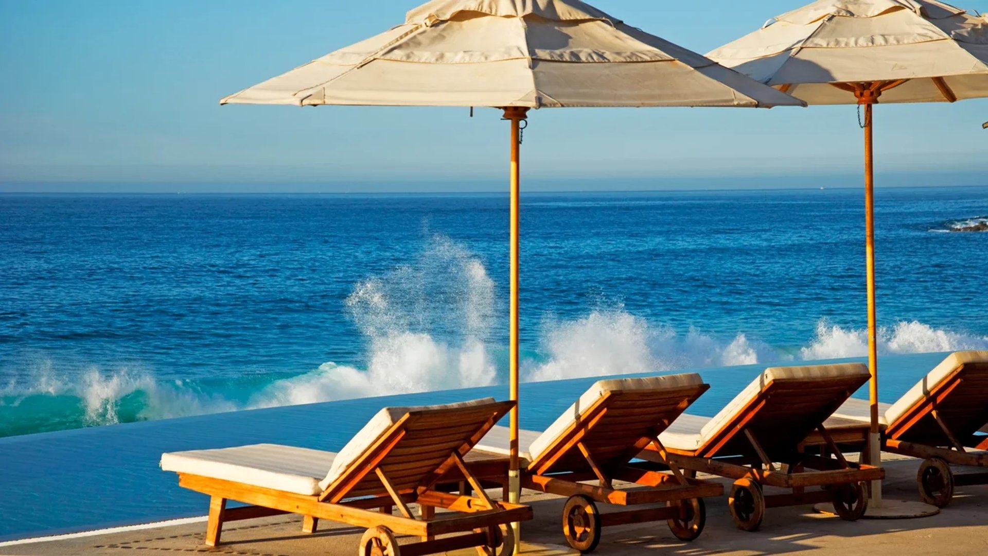 Lounge chairs under beige umbrellas on a clifftop overlooking turquoise ocean waves