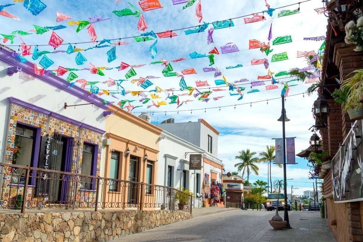 Colorful papel picado banners strung across a sunny colonial street with colonial buildings and palm trees in the background